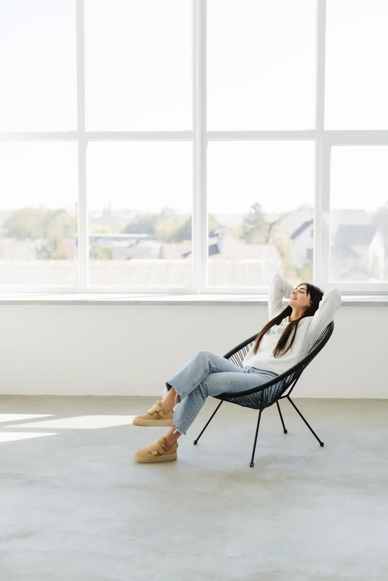 Völlige Entspannung: Eine attraktive junge Frau hält die Hände hinter dem Kopf und lächelt, während sie zu Hause auf einem Stuhl sitzt.